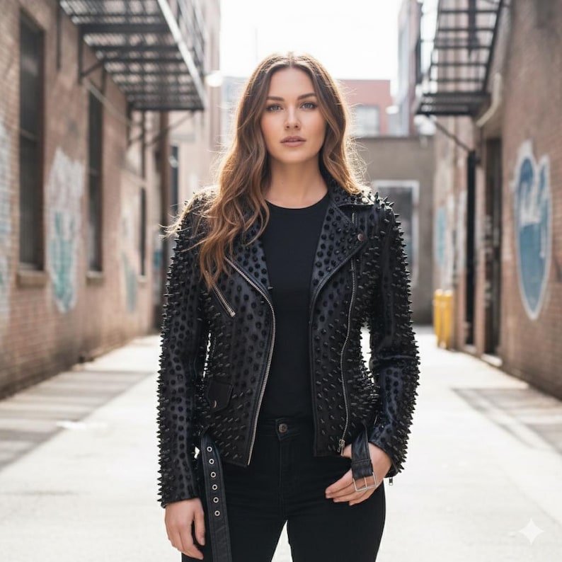May include: A black leather motorcycle jacket featuring an all-over pattern of small, pointed black studs, a notched lapel, silver-toned metal zippers, and an adjustable buckled belt at the waist, worn over a plain black shirt and black jeans.