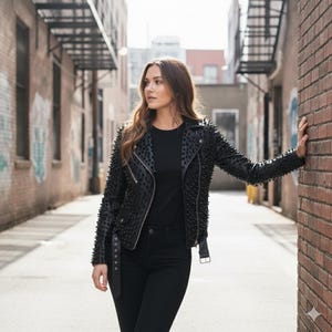 May include: A black leather motorcycle jacket featuring dense, uniform stud embellishments across the sleeves and front panels, paired with a plain black t-shirt and black denim jeans. The jacket includes a silver-toned zipper and a buckled belt.
