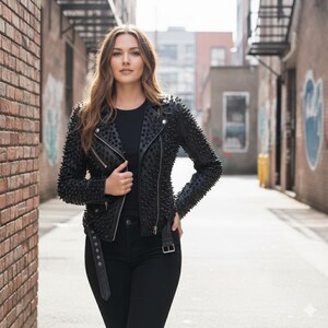 May include: A black leather jacket featuring an all-over spiked texture, silver-toned metal zippers, and a buckled belt at the waist. The jacket is worn over a plain black shirt and paired with black pants, creating a monochromatic ensemble.