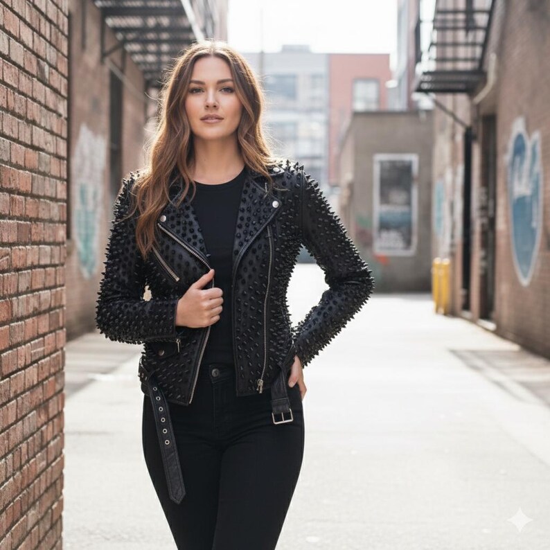 May include: A black leather jacket featuring an all-over spiked texture, silver-toned metal zippers, and a buckled belt at the waist. The jacket is worn over a plain black shirt and paired with black pants, creating a monochromatic ensemble.