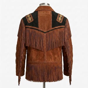 K&ouml;nnte beinhalten: Eine braune Wildlederjacke mit langen Fransen am R&uuml;cken und an den &Auml;rmeln. Das Kleidungsst&uuml;ck verf&uuml;gt &uuml;ber schwarze Wildledereins&auml;tze an den Schultern mit dekorativen kreisf&ouml;rmigen Stickereien, geflochtenen Lederverzierungen und einer R&uuml;ckenpasse.