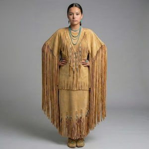 May include: A woman in a tan suede fringed dress and shawl, with turquoise jewellery. The dress has detailed fringe on the sleeves, bodice, and skirt, creating a flowing silhouette. The outfit is completed with matching moccasins.