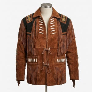 K&ouml;nnte beinhalten: Eine braune Wildlederjacke mit langen Fransen an Brust und &Auml;rmeln. Das Kleidungsst&uuml;ck verf&uuml;gt &uuml;ber schwarze Wildledereins&auml;tze an den Schultern mit dekorativen Stickereien, wei&szlig;en knochenartigen Akzenten und zwei Bindeb&auml;ndern vorne.