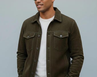 May include: An olive green button-up jacket with a pointed collar, long sleeves, and two chest pockets with buttoned flaps. The jacket is worn over a plain white crew neck shirt. The garment features a smooth texture and dark buttons.