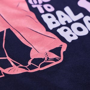 以下が含まれることがあります： ダンサーの足と靴のピンク色のグラフィックが描かれた、濃いネイビーブルーのTシャツのクローズアップ。 「TO BALBOA！」の文字が白とピンクでプリントされています。 デザインは、ダンス愛好家向けである可能性があります。