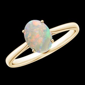 May include: A gold ring featuring an oval opal gemstone. The opal displays a play of color with iridescent hues of green, orange, and pink. The ring has a simple, elegant design with four prongs securing the gemstone.