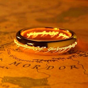 Op de afbeelding: Een goudkleurige ring met een oranje gloed aan de binnenkant, versierd met witte letters. De ring ligt op een kaart met het woord "MORDOR" zichtbaar. Het ontwerp is geïnspireerd op een fantasiethema.