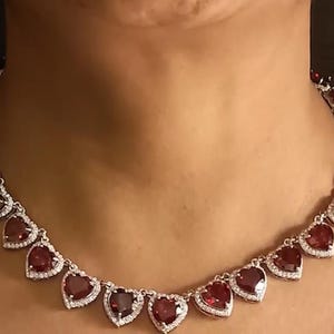 Könnte beinhalten: Eine silberne Halskette mit herzförmigen, roten Edelsteinen, die jeweils von kleinen, klaren Steinen umgeben sind. Die Halskette wird um den Hals einer Person getragen und zeigt ihr Design und ihre Farbe.