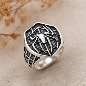 Pode incluir: Um anel de tom prateado com uma face hexagonal apresentando um emblema de aranha em relevo centrado sobre um padrão de teia texturizado. A banda e as laterais do anel também exibem um design de teia em grade com detalhes oxidados escuros.