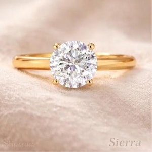 Può includere: Un anello d'oro con una grande gemma rotonda e trasparente. L'anello ha una fascia semplice e quattro griffe che tengono la gemma. L'anello è posto su uno sfondo morbido e chiaro. La parola "Sierra" è visibile.