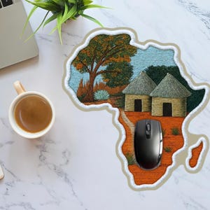 May include: An African-themed mousepad featuring an embroidered map of the continent with a landscape scene. The scene includes trees, huts, and a black computer mouse. A laptop, coffee cup, notebook, and pencil are also visible.