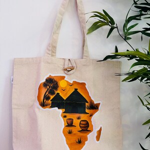 Puede incluir: Bolso tote de lona beige con cierre de palanca de madera. El bolso presenta una vibrante ilustración naranja y negra del continente africano, con cabañas, árboles y un barril. Tiene asas largas.