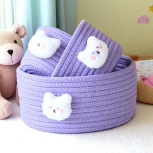 May include: Three lavender rope baskets, each featuring a white bear applique with pink cheeks and a brown nose. The baskets are nested together, offering a decorative storage solution. The baskets are ideal for storing toys or other items.