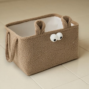 Puede incluir: Una cesta de almacenamiento rectangular marrón con un exterior esponjoso y texturizado. La cesta tiene dos ojos blancos saltones y dos pequeñas asas en forma de oreja. El interior es blanco y hay dos asas más grandes en los laterales.