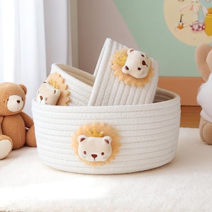 May include: Three white rope baskets, each decorated with a lion face applique. The largest basket contains the smaller ones. The lion appliques feature a yellow mane and a small gold crown. These baskets are suitable for nursery storage.