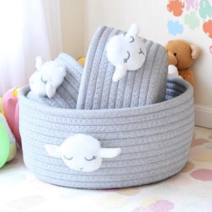 May include: A grey, round woven storage basket with white sheep appliqués. The basket contains folded grey blankets and additional sheep decorations. The basket is designed for nursery or children's room storage.