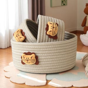 Pode incluir: Um conjunto de três cestos de armazenamento cinza claro, um grande e dois menores, aninhados. Cada cesto é adornado com uma aplicação de feltro de leão. Para o quarto das crianças.