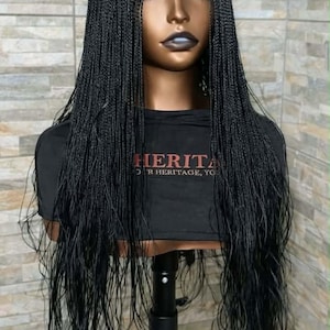 May include: A black braided wig on a mannequin head. The wig features long, thin braids and a lace front. The mannequin is wearing a black t-shirt with the word "HERITAGE" printed on it. The background is a light-colored brick wall.