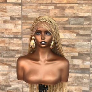 May include: A mannequin head with a dark skin tone wearing a long, blonde braided wig. The wig features fine, micro-braided strands and a lace front hairline. The mannequin is adorned with large, gold-toned, teardrop-shaped earrings.