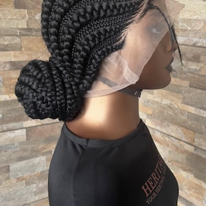 May include: A black braided wig styled in a bun. The wig features a lace front and is worn on a mannequin. The mannequin is wearing a black shirt with the text "HERITAGE YOUR HERITAGE" in red.