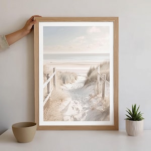 Puede incluir: Una impresión enmarcada de una escena de playa con un sendero arenoso que conduce al océano. La impresión está en un marco de madera clara y presenta un paisaje costero sereno con dunas de arena y un cielo despejado. Una pequeña suculenta en una maceta blanca está sobre la mesa.