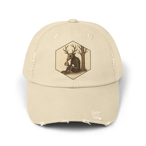 May include: Beige distressed baseball cap featuring a hexagonal graphic of a deer-like creature with antlers, seated near a tree. The illustration is in shades of brown and tan. The cap has a curved brim and distressed detailing.