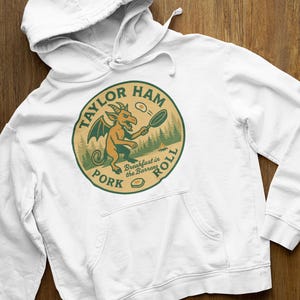 Puede incluir: Sudadera con capucha blanca con bolsillo delantero y cordón. La parte delantera presenta un diseño gráfico circular con el texto "TAYLOR HAM PORK ROLL". En el interior, un dragón de dibujos animados cocina el desayuno.