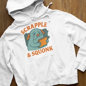 Può includere: Felpa con cappuccio bianca con tasca frontale e coulisse. La felpa presenta un'illustrazione a cartoni animati di una creatura turchese che tiene in mano un pezzo di pane, con la scritta "SCRAPPLE & SQUONK" in arancione.