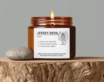 May include: A lit, brown glass jar candle with a label that reads "JERSEY DEVIL." The label includes a graphic of a devil and the phrases "Doesn't do small talk," "Always down for chaos," and "Loves bonfire nights." A grey stone sits next to the candle.