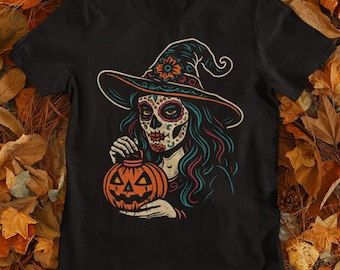 May include: Black t-shirt with a colourful illustration of a sugar skull witch holding a carved pumpkin. The witch wears a pointed hat with a floral design and has teal hair. The pumpkin is orange with a carved face.