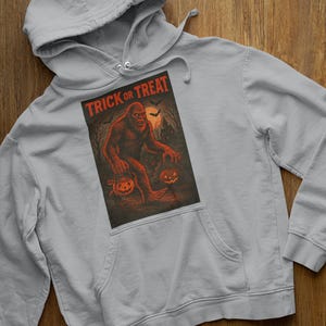 May include: A pale grey hoodie with a Halloween graphic. The design shows a Bigfoot-like figure with a jack-o'-lantern and a trick-or-treat bucket, with the words "TRICK or TREAT" above. The graphic uses orange and black colours.