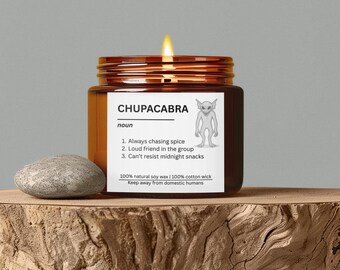 May include: A lit soy wax candle in a brown glass jar with the word "CHUPACABRA" and a definition printed on the label. A small gray stone sits next to the candle on a wooden surface.
