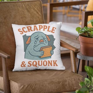 May include: White square throw pillow with a cartoon illustration of a sad, blue creature holding a piece of toast. The pillow has the words "SCRAPPLE & SQUONK" in orange lettering.