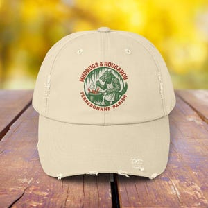 May include: A distressed beige baseball cap with a circular graphic featuring a wolf-like creature cooking over a pot, with the text "MUDBUGS & ROUGAROU TERREBONNE PARISH" in a circular design. The cap has a curved brim.