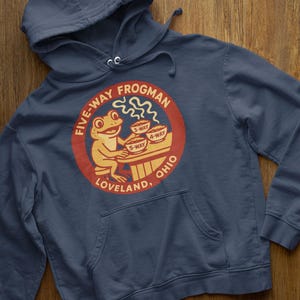 May include: Navy blue hooded sweatshirt featuring a circular graphic with a frog character. The graphic reads "FIVE-WAY FROGMAN" and includes text for "3-WAY", "4-WAY", and "5-WAY" options, with "LOVELAND, OHIO" below. The sweatshirt has a front pocket.