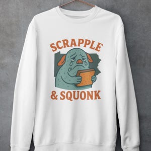 May include: White sweatshirt with the words "SCRAPPLE & SQUONK" in orange and green. The graphic features a sad, crying cartoon character holding a piece of toast. The character is light blue with orange ears.