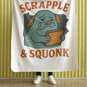 Può includere: Coperta bianca con la scritta "SCRAPPLE & SQUONK" in arancione. La coperta presenta un personaggio dei cartoni animati con la pelle turchese, le orecchie arancioni e un'espressione triste, che tiene in mano un oggetto marrone.