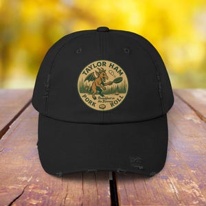 Puede incluir: Gorra de béisbol negra con ala desgastada. La gorra presenta un parche circular con el texto "TAYLOR HAM PORK ROLL" alrededor de un dragón de dibujos animados cocinando en una sartén. El fondo es una escena de bosque.