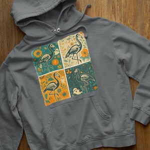 Peut inclure: Un sweat à capuche gris avec un motif à quatre panneaux représentant une illustration de squelette de flamant rose. Chaque panneau a un arrière-plan différent avec des citrouilles, des fleurs et des fantômes. Le sweat a une poche avant et une capuche à cordon.