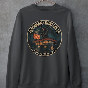 May include: Dark gray sweatshirt featuring a circular graphic with the text "Mothman & Roni Rolls" and "Point Pleasant, WV." The graphic depicts a creature with wings and a diner. The sweatshirt is made of a soft material.