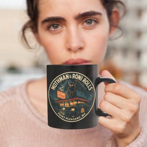 May include: Black ceramic mug featuring a graphic of a Mothman creature with the text "Mothman & Roni Rolls" and "Point Pleasant, WV". The design includes a diner and a dark, ominous sky.