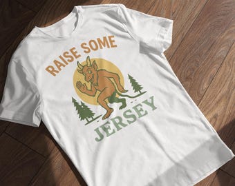 May include: White t-shirt with a cartoon illustration of a horned creature running, set against a yellow circle and two green pine trees. The text "RAISE SOME JERSEY" is printed above and below the graphic in brown and green.
