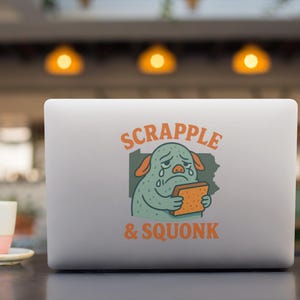 Può includere: Un laptop argentato con un adesivo raffigurante un mostro verde che piange e tiene in mano un toast. Il testo sull'adesivo recita "SCRAPPLE & SQUONK" in arancione. Una tazza di caffè è in primo piano.