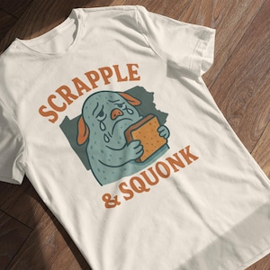May include: White t-shirt with a distressed graphic of a crying blue creature holding a piece of bread. The text "SCRAPPLE & SQUONK" is printed above and below the illustration in orange.