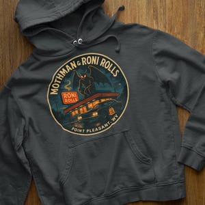 May include: Dark gray hooded sweatshirt with a graphic featuring a winged creature perched atop a diner. The design includes the text "Mothman & Roni Rolls" and "Point Pleasant, WV" in a circular emblem. The sweatshirt has a front pocket.