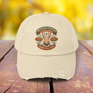 May include: A distressed beige baseball cap with a graphic featuring a jackalope and the text "Mountain Oysters & Jackalopes Frontier Fare" in a circular design. The cap has a curved brim and a worn appearance.