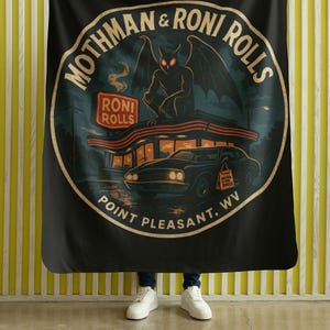 May include: Black blanket featuring a circular design with the text "Mothman & Roni Rolls, Point Pleasant, WV." The design includes a creature with wings, a diner, and a car. The blanket is held up by a person.
