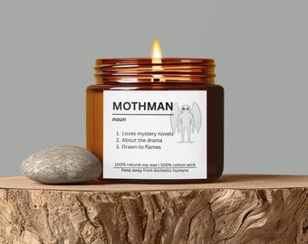 May include: A lit candle in a brown glass jar with the word "MOTHMAN" on the label. The label also includes a cartoon illustration of a Mothman and the phrases "Loves mystery novels", "About the drama", and "Drawn to flames". A gray stone sits to the left.