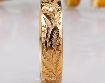 May include: A gold ring with an engraved floral pattern. The ring has detailed carvings of leaves and flowers. The brand name "CARATEVARA" is visible. The background features yellow flowers.