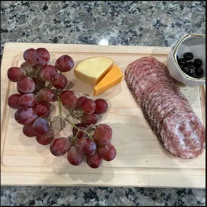 May include: A rectangular light-coloured wooden serving board holds a large cluster of red grapes, two wedges of yellow and orange cheese, a long row of thinly sliced salami with a peppered edge, and a small white bowl filled with black olives.